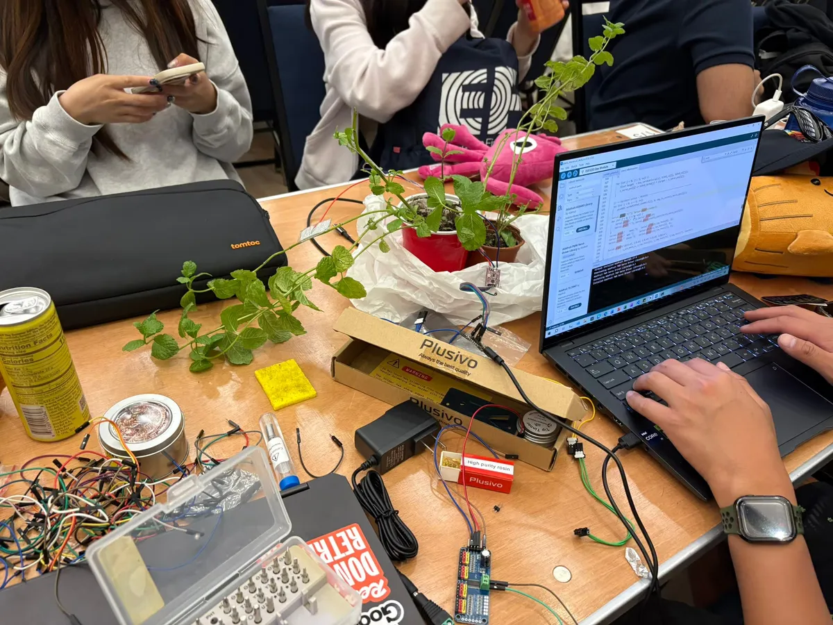 A busy hackathon table with the Plantcasso plant, laptop, electronics, and tools