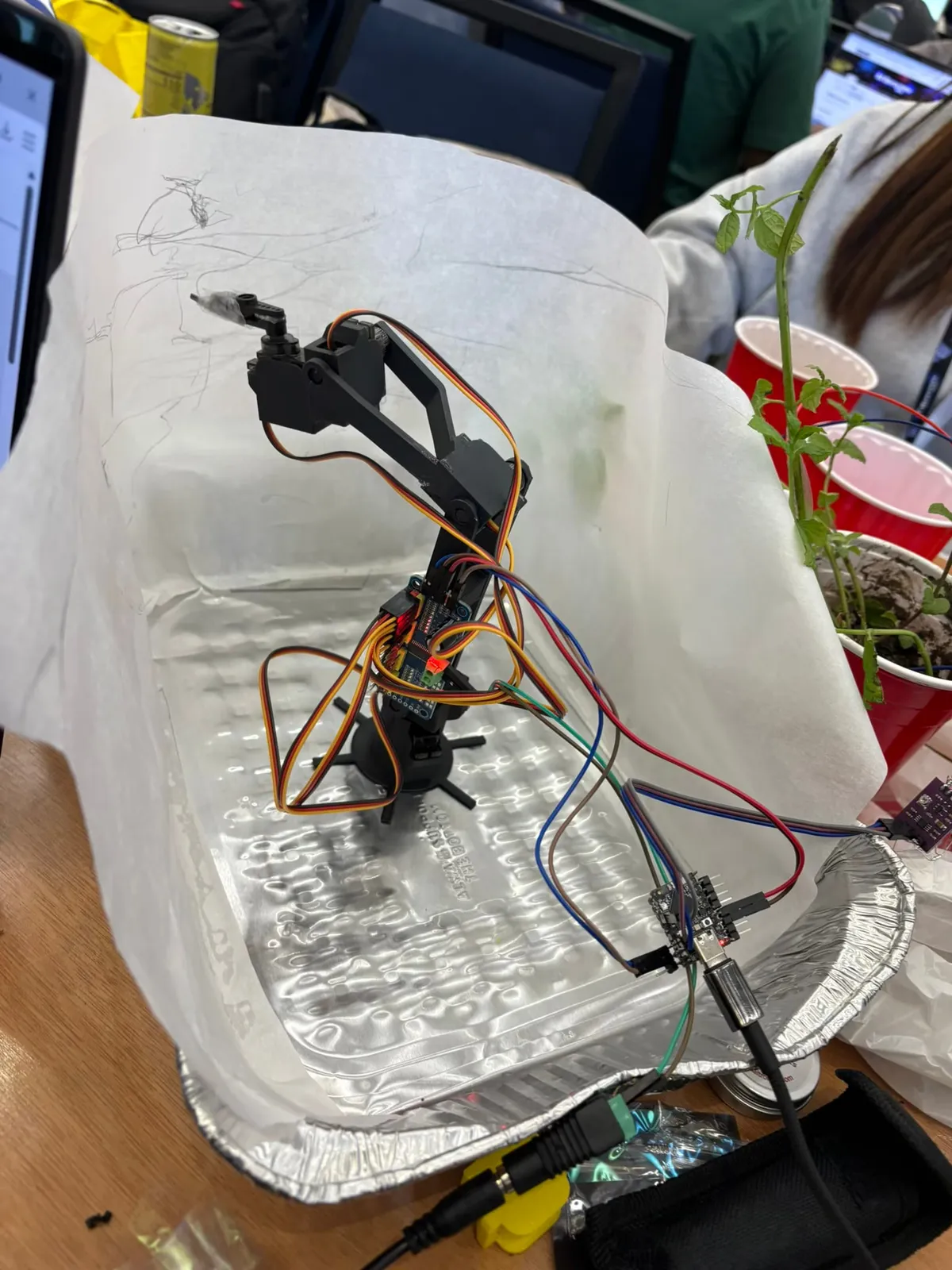 A small robot arm wired inside a foil enclosure with a mint plant beside it